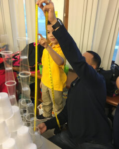 A teacher and a student are next to a tower of plastic cups. The teacher is holding an expanded measuring tape, and the student is indicating how tall the tower is.